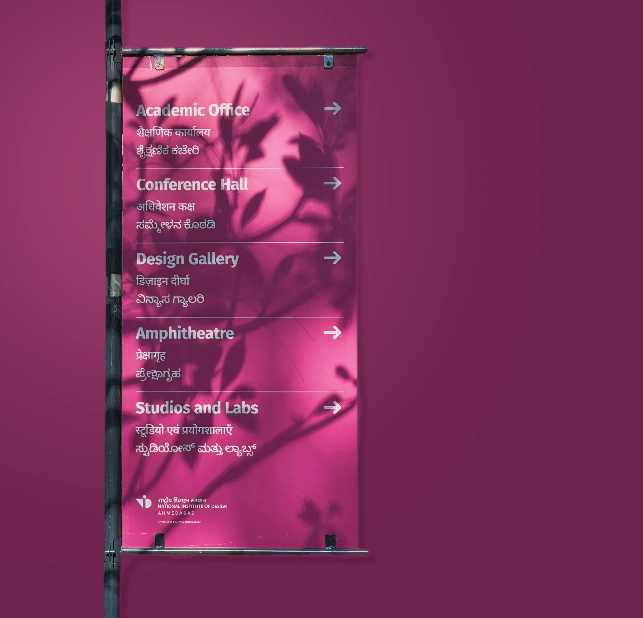 Pink hanging banner sign listing Academic Office, Conference Hall, Design Gallery, Amphitheatre and Studios and Labs in English, Hindi and Kannada